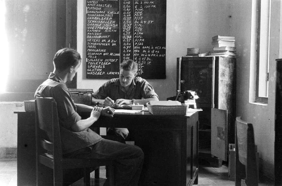 Een sergeant achter een bureau noteert, voor hem zit een soldaat. Achter de serg, Bestanddeelnr 369