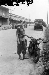 Een sergeant kijkt naar de fotograaf, achter hem loodsen en vrachtwagen (Tandjon, Bestanddeelnr 5969