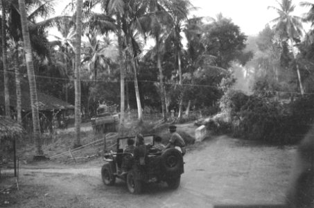 Een tank omzijlt een omgehakte boom en rijdt een kampong in Op de voorgrond ee…, Bestanddeelnr 333-3-3