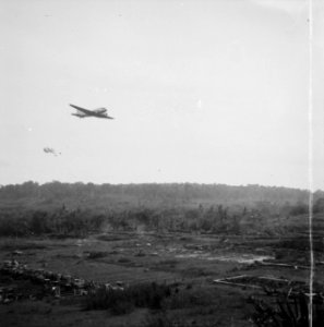 Een vliegtuig werpt goederen per parachute af over een terrein waarop gebouwen , Bestanddeelnr 13053