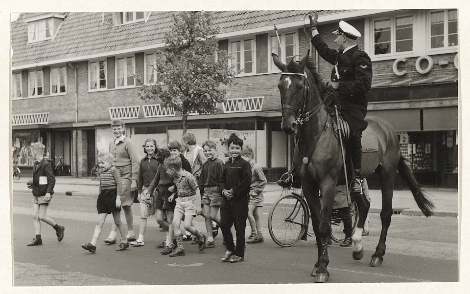 Een agent te paard brengt schoolgaande kinderen veilig over de gevaarlijke Rijksstraatweg naar de Vergierdeweg. NL-HlmNHA 54004610