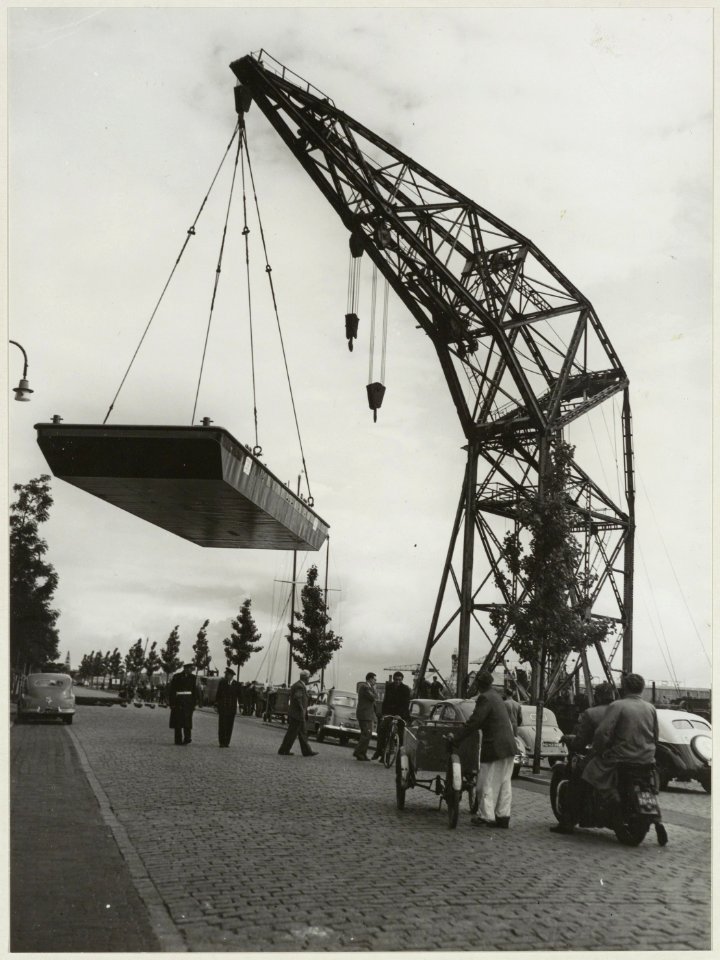 Een 30.000 kg zware scheepsromp bungelt aan de machtige arm van de drijvende bok ( hijskraan) van Conrad Stork aan het Spaarne nabij de Floresstraat. NL-HlmNHA 54010641