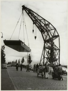 Een 30.000 kg zware scheepsromp bungelt aan de machtige arm van de drijvende bok ( hijskraan) van Conrad Stork aan het Spaarne nabij de Floresstraat. NL-HlmNHA 54010641