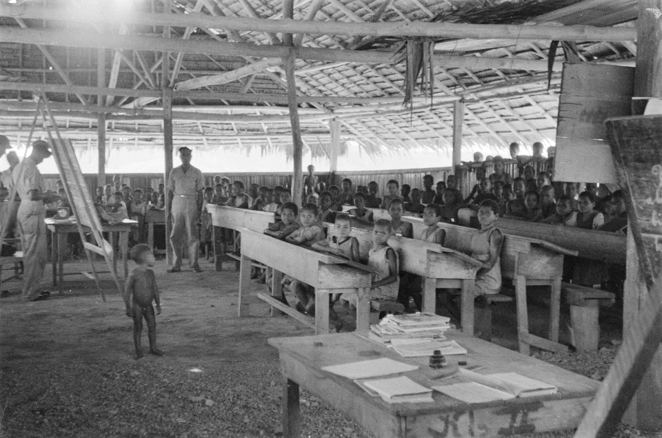 Een grote groep schoolkinderen zit achter schoolmeubilair in een schoolgebouw, Bestanddeelnr 440-6-5