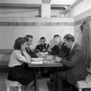 Een groep mensen, waaronder een vrouw en een marechaussee rond een tafel in een, Bestanddeelnr 900-4486
