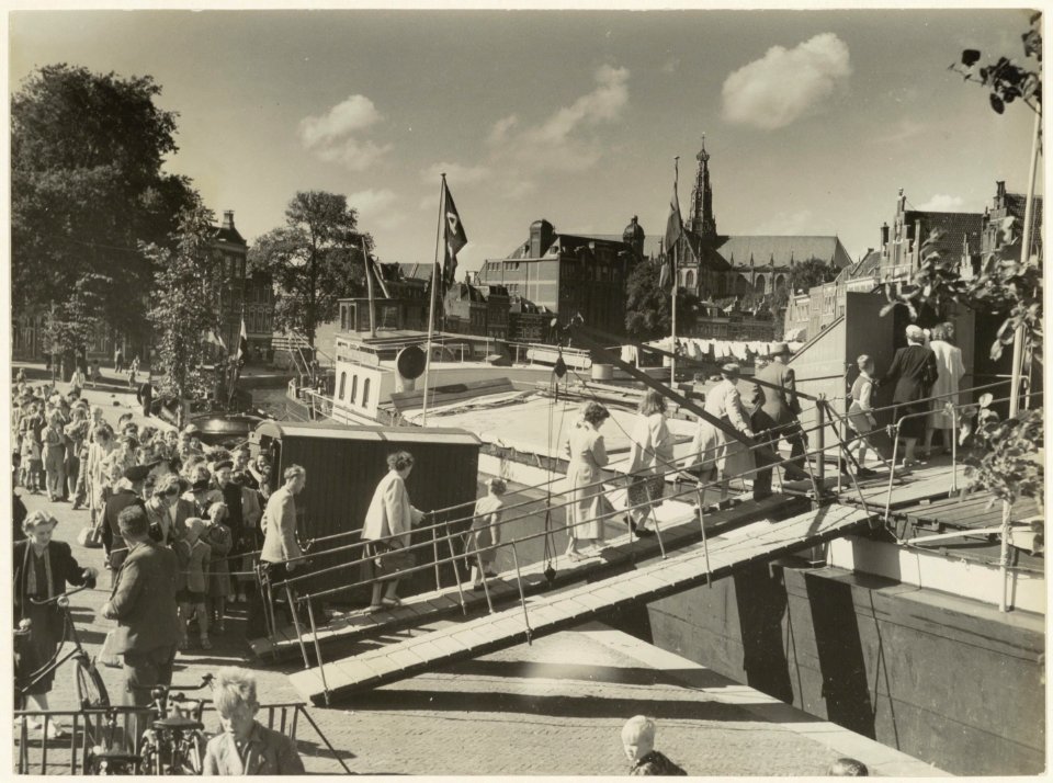 Een grote groep mensen betreedt via de loopplank een afgemeerd schip in het Spaarne. NL-HlmNHA 54011291