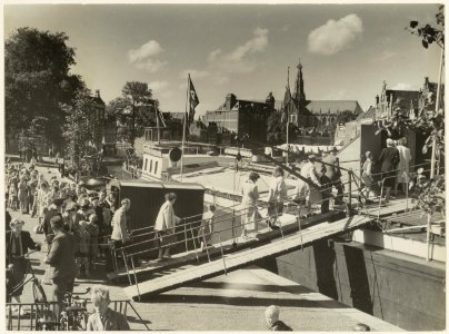 Een grote groep mensen betreedt via de loopplank een afgemeerd schip in het Spaarne. NL-HlmNHA 54011291