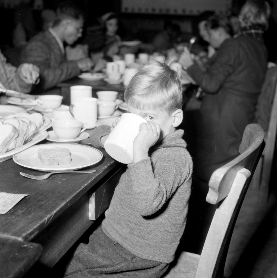 Een jongetje zit aan een eettafel met veel mensen en drinkt uit beker, Bestanddeelnr 255-9480