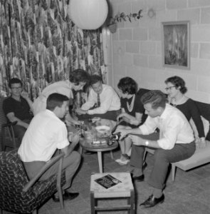 Gasten op een feestje in huiselijke kring rond de tafel met een hapje en een dra, Bestanddeelnr 255-4320