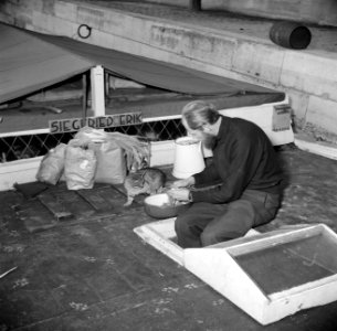 Gezeten aan dek van een schip met zijn benen in een luik, snijdt een man de groe, Bestanddeelnr 252-9400