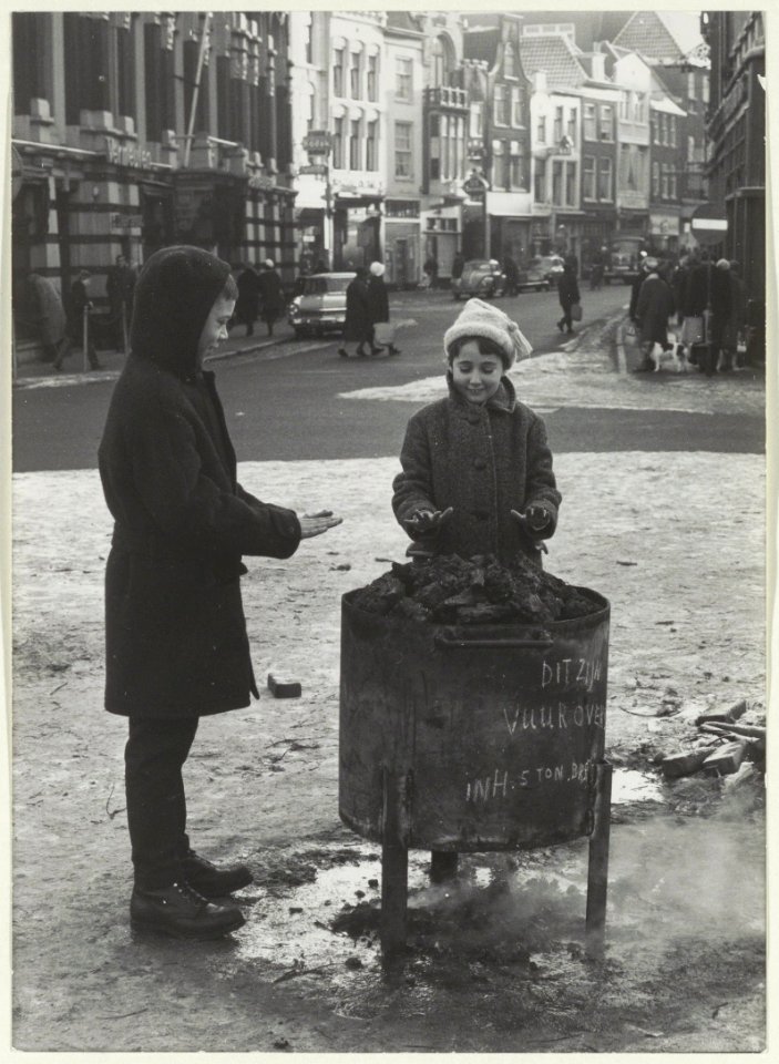 Handen warmen aan een vuuroven in de sneeuw, op de Grote Markt ziende in de Grote Houtstraat. NL-HlmNHA 54008967