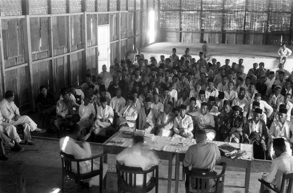 Groot gezelschap Indonesische mannen (met peci) zitten in een grote zaal. Op het, Bestanddeelnr 3357