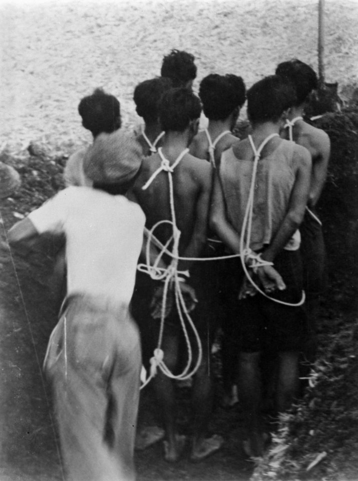 Groep mannen, met touwen geboeid staan in een greppel. Achter hen een man met li, Bestanddeelnr 8807