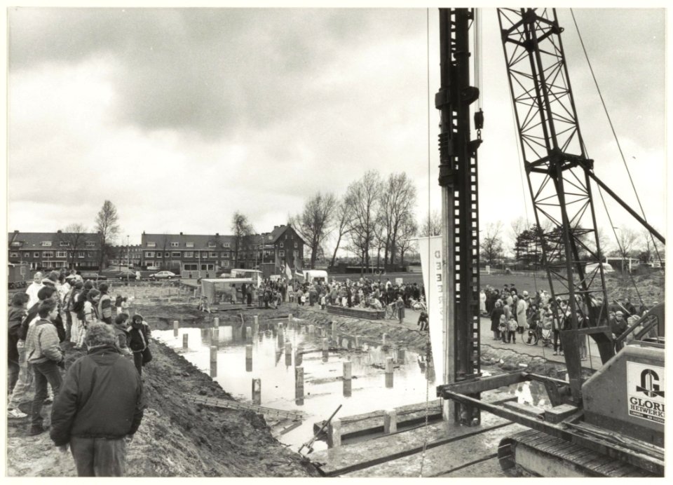 Het slaan van de eerste paal voor de nieuwbouw aan het Nieuw Guineaplein, ziende naar het noordoosten. De huizen op de achtergrond staan aan de Jan Gijzenkade, hoek Vondelweg. NL-HlmNHA 54023728