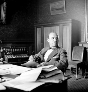 Heer met vlinderstrik zittend achter bureau met rechts van hem een kleine telefo, Bestanddeelnr 255-8800