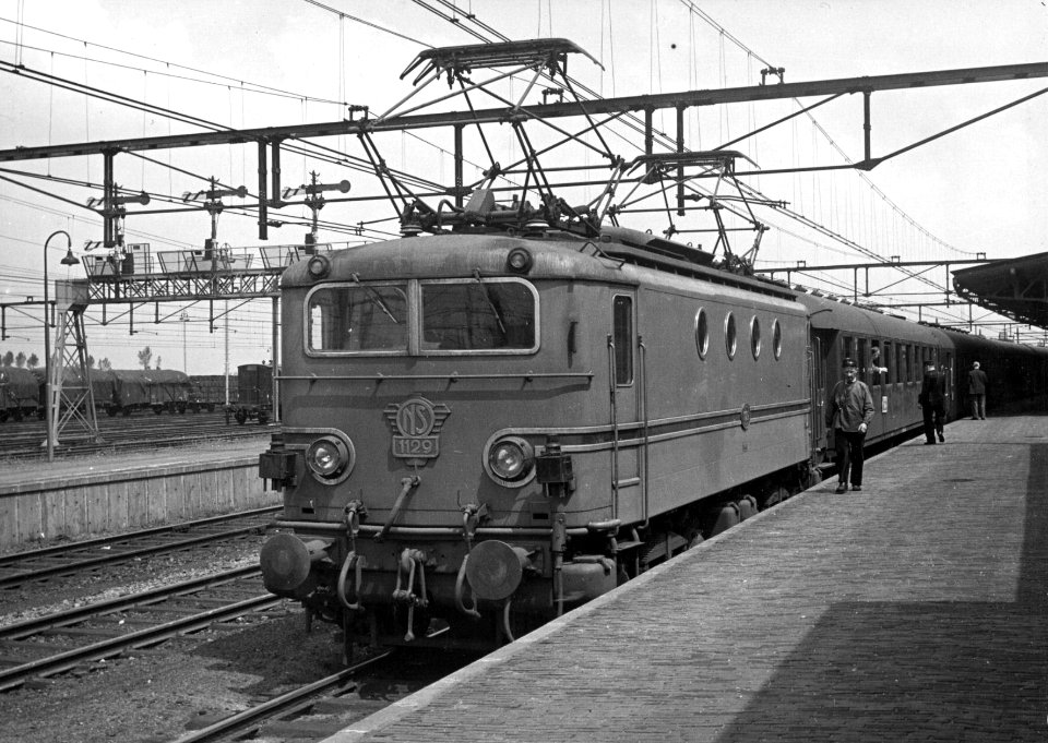 HUA-154998-Afbeelding van de electrische locomotief nr. 1129 (serie 1100) van de N.S. met rijtuigen langs het perron van het N.S.-station Roosendaal te Roosendaal
