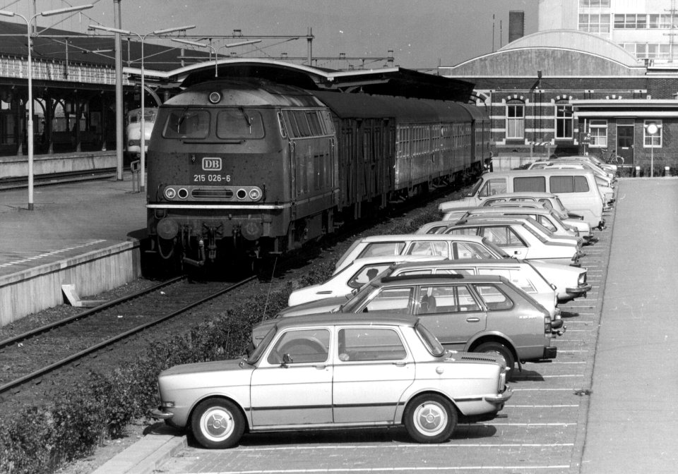 HUA-154560-Afbeelding van de diesellocomotief 215 026-6 van de D.B. met rijtuigen (trein naar Kleve) langs het perron van het N.S.-station Nijmegen te Nijmegen. Rechts een deel van de P+R-parkeerplaats