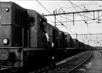 HUA-154311-Afbeelding van twee diesel-electrische locomotieven uit de serie 2400-2500 (voorop nr. 2491) van de N.S. met een goederentrein te Roosendaal. De machinist krijgt de vrachtbrieven aangereikt