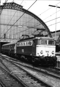 HUA-151130-Afbeelding van de electrische locomotief nr. 1113 (serie 1100) van de N.S. met intenationale rijtuigen langs het perron van het N.S.-station Amsterdam C.S. te Amsterdam