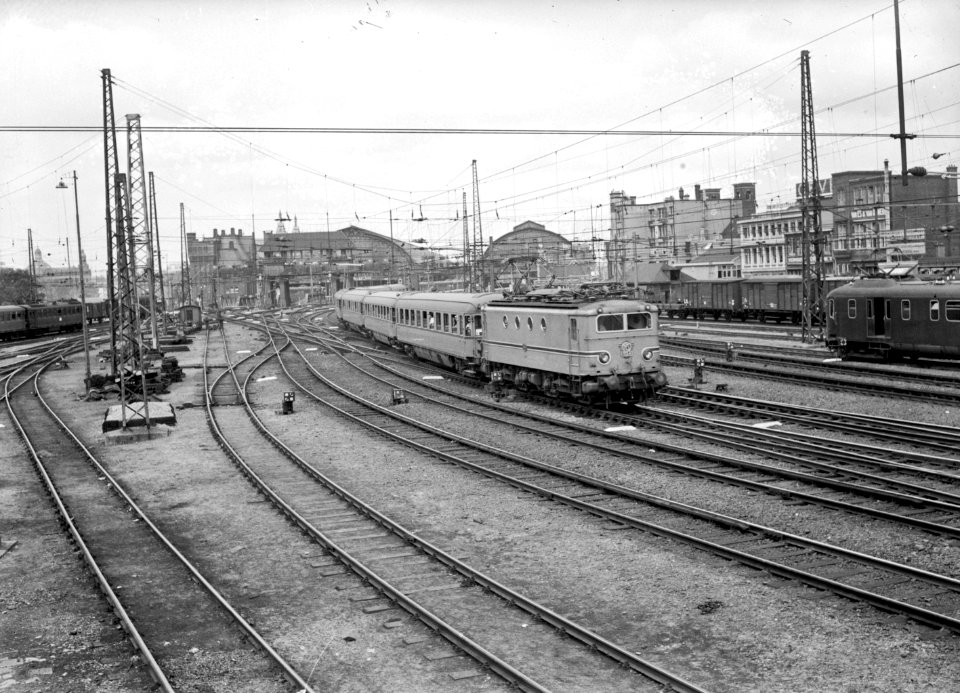 HUA-151161-Afbeelding van de electrische locomotief nr. 1134 (serie 1100) van de N.S. met plan D-rijtuigen in turkooise beschildering op het emplacement aan de oostzijde van het N.S.-station Amsterdam C.S. te Am