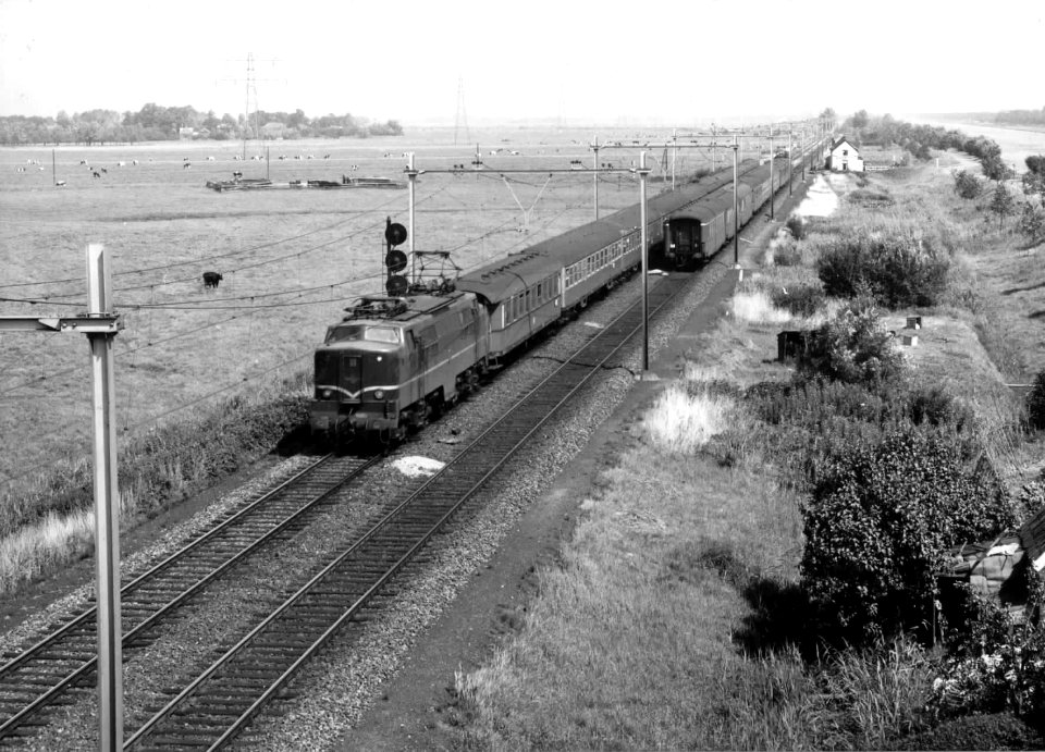 HUA-150937-Afbeelding van de electrische locomotief nr. 1202 (serie 1200) van de N.S. met rijtuigen (tijdens passeren van een tegentrein) in een weidelandschap ter hoogte van Loenen aan de Vecht. Op de achtergro