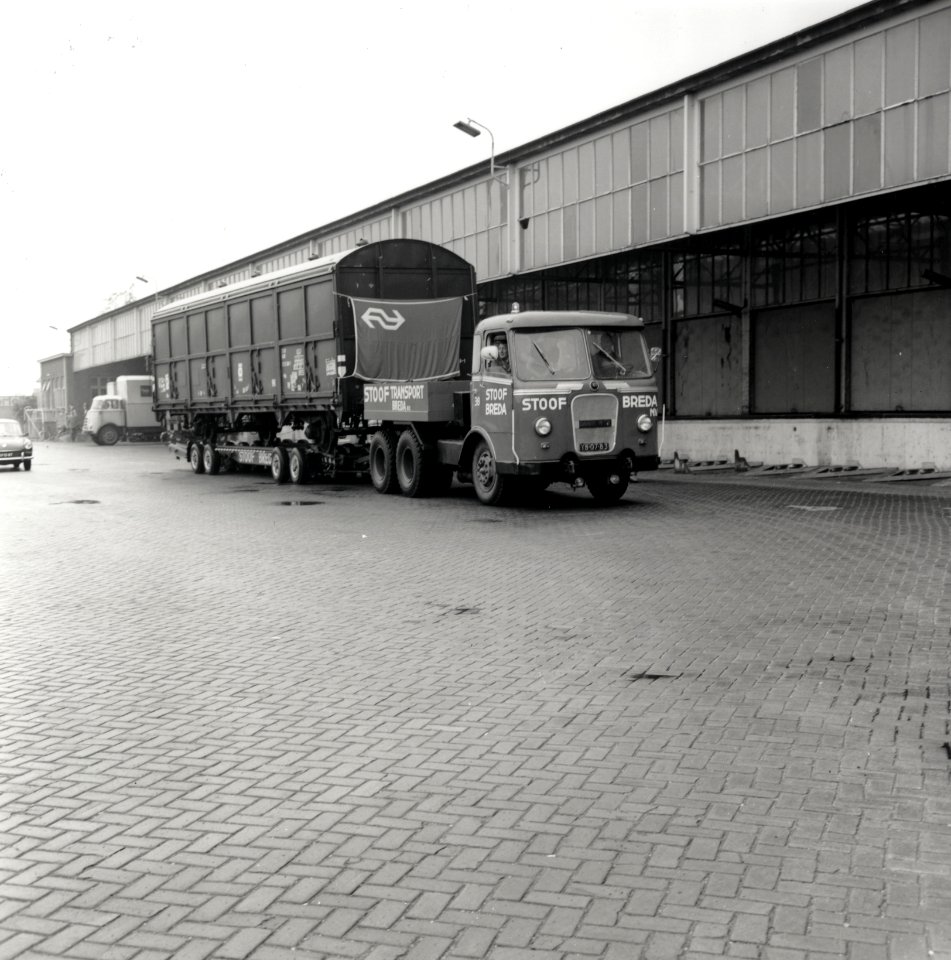 HUA-168029-Afbeelding van het transport van een goederenwagen type Hbis van de N.S. per vrachtauto ("rolwagenvervoer" of "strassenroller") door de firma Stoof te Breda. Op de achtergrond de goederenloods van Van