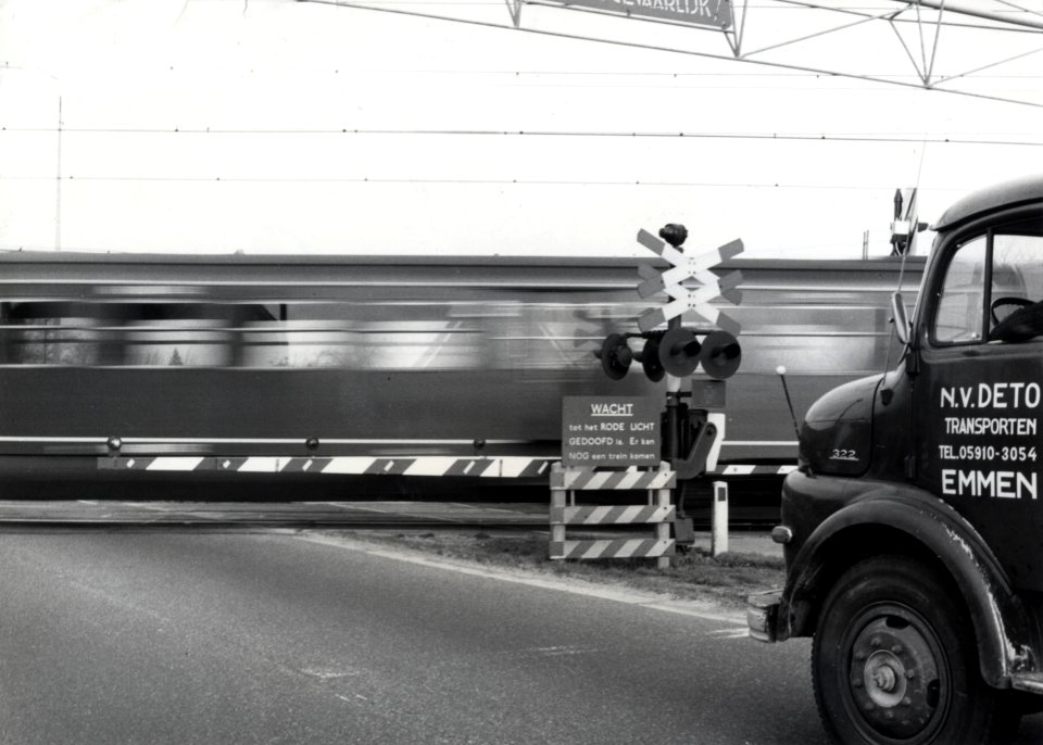 HUA-170532-Afbeelding van een AHOB bij een spoorwegovergang, tijdens het passeren van een trein, met rechts een gedeelte van een vrachtauto van de N.V. DETO te Emmen