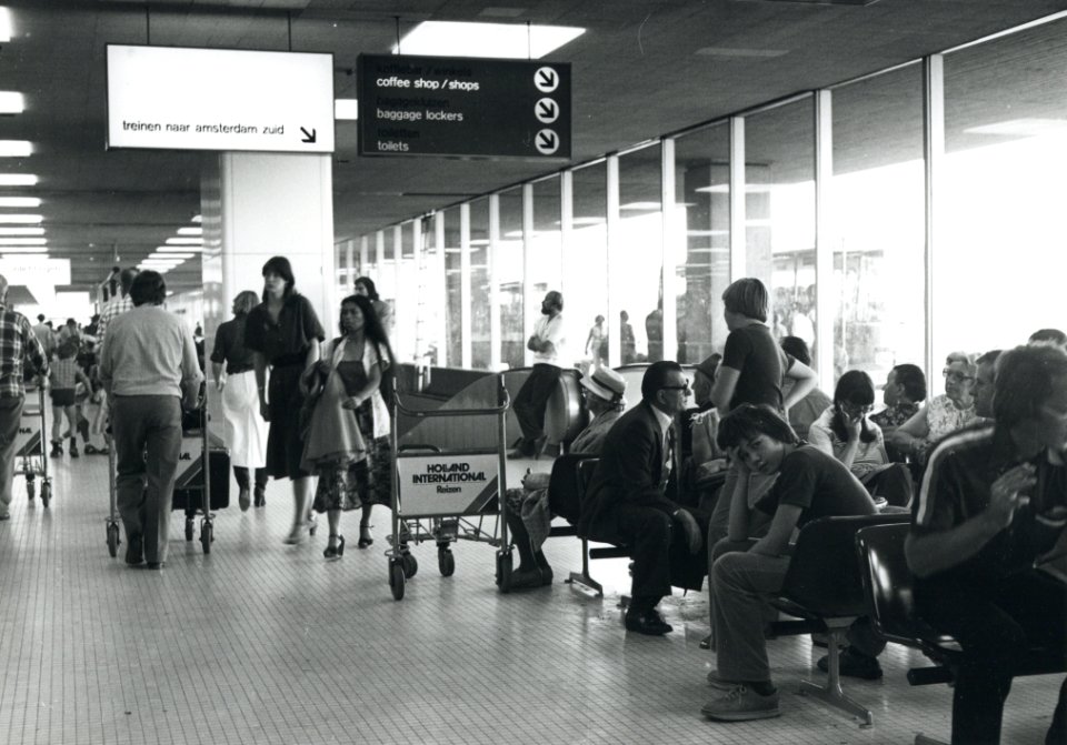 HUA-170845-Interieur van één van de vertrekhallen van de luchthaven Schiphol, met wachtende luchtreizigers en linksboven een verwijzing naar de treinen naar Amsterdam Zuid