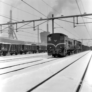 HUA-159802-Afbeelding van twee diesel-electrische locomotieven uit de serie 2200-2300 van de N.S. (voorop nr. 2334) met een kolentrein op het besneeuwde emplacement te Heerlen. Op de achtergrond de Oranje Nassau