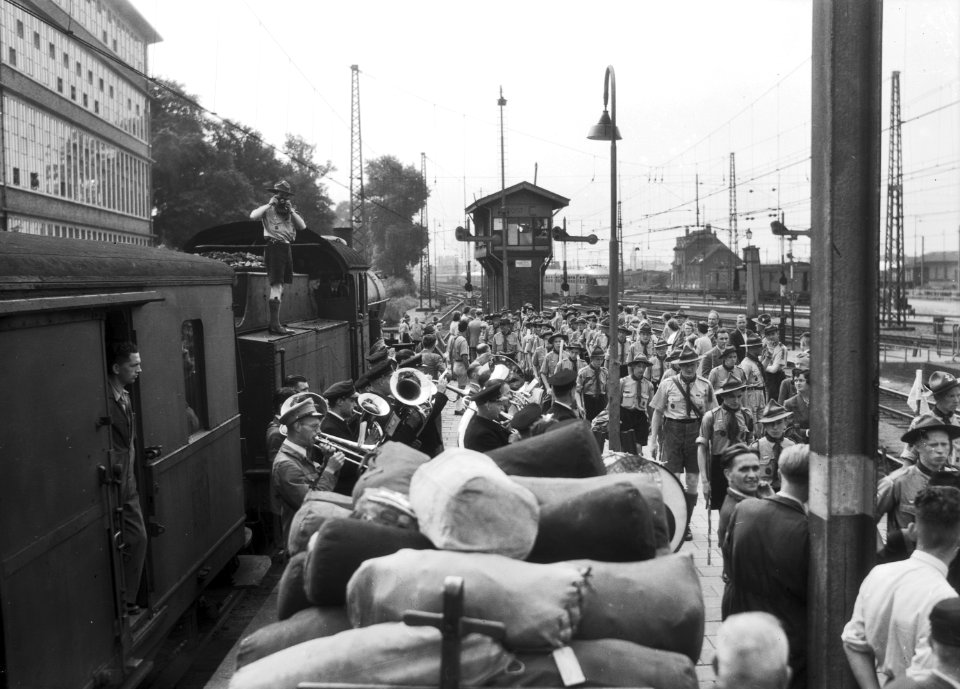 HUA-167546-Afbeelding van het vertrek van een groep padvinders per trein naar de Jamboree te Oostenrijk op het perron van het N.S.-station Utrecht C.S. te Utrecht, met op de voorgrond een harmonieorkest, links e