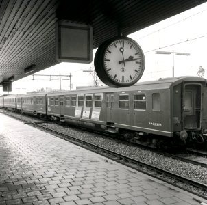 HUA-167742-Afbeelding van de stationsklok op het eerste perron van het N.S.-station Driebergen-Zeist te Driebergen-Rijsenburg, met op de achtergrond een getrokken trein bestaande uit rijtuigen plan E en plan K