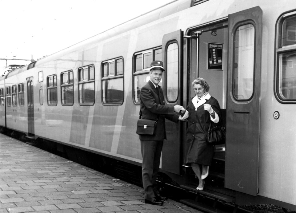 HUA-165773-Afbeelding van een conducteur van de NS tijdens het helpen bij het uitstappen van een dame op leeftijd uit een electrisch treinstel plan V mat 1964 
