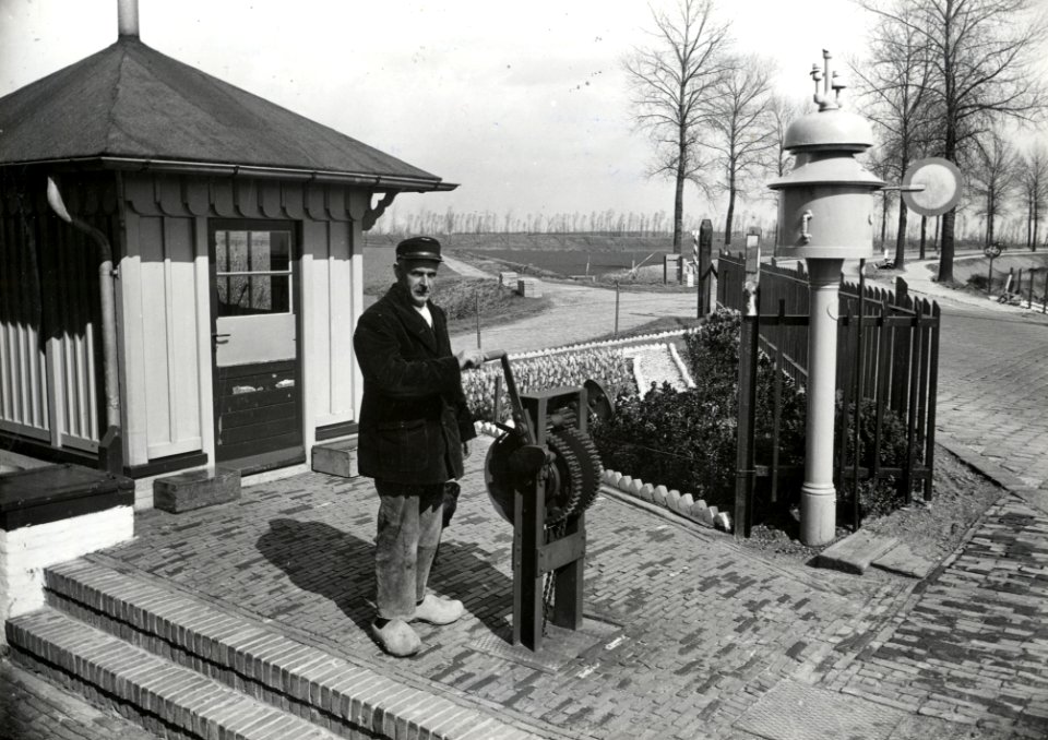 HUA-169411-Afbeelding van overwegwachter G.D. van de Broek bij het mechanisme voor de bediening van de overwegbomen bij wachtpost 24a aan de Oude Rijksweg te Krabbendijke, met rechts het kloksein