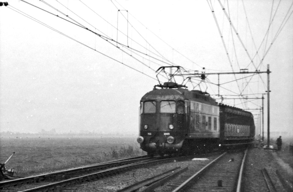 HUA-164303-Afbeelding van de electrische locomotief nr. 1003 (serie 1000) van de N.S. met een trein op een overloopwissel ter hoogte van Abcoude, tijdens de werkzaamheden ten behoeve van de baanverzwaring tussen