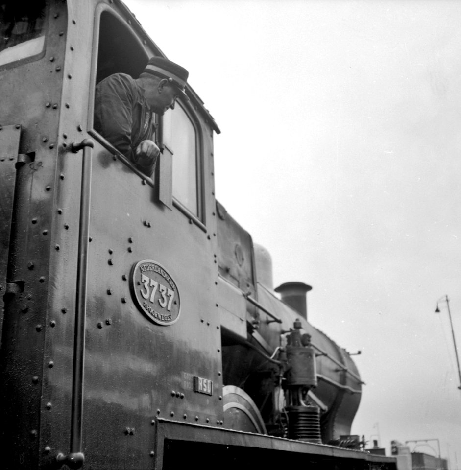 HUA-153437-Afbeelding van de machinist hangende uit het raam van de stoomlocomotief nr 3737 serie 3701 3815 van de NS tijdens zijn laatste rit
