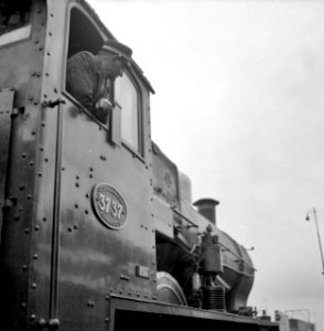 HUA-153437-Afbeelding van de machinist hangende uit het raam van de stoomlocomotief nr 3737 serie 3701 3815 van de NS tijdens zijn laatste rit