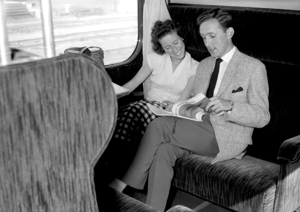 HUA-152859-Afbeelding van twee reizigers in een trein, met het tijdschrift "Tussen de Rails" van de N.S