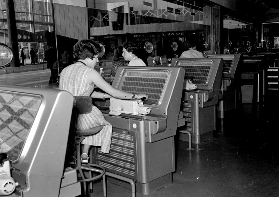 HUA-152564-Afbeelding van lokettisten in het plaatskaartenkantoor in het NS station Rotterdam CS te Rotterdam met enkele lokettistes achter de AEG plaatskaartend