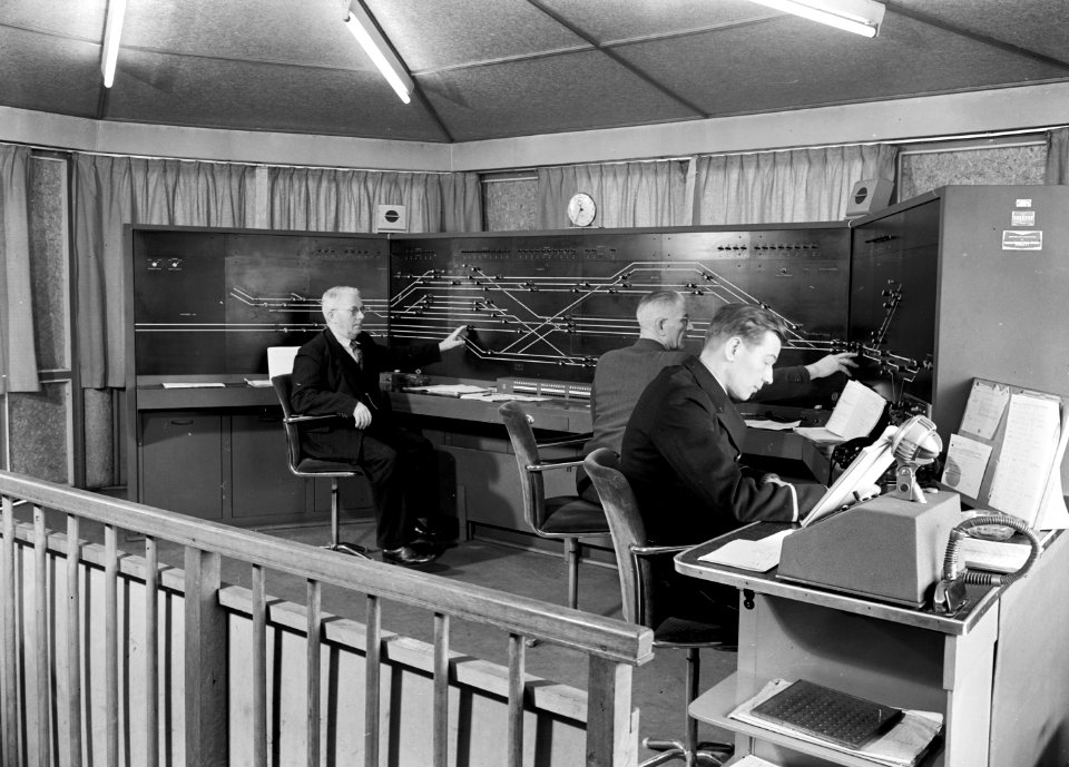 HUA-159667-Interieur van het seinhuis bij het N.S.-station Arnhem te Arnhem met de treindienstleiders bij het NX-bedieningstableau