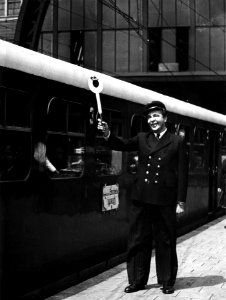 HUA-163922-Afbeelding van Wim Kan van het ABC-Cabaret als perronopzichter op het perron van het N.S.-station Amsterdam C.S. te Amsterdam, tijdens het geven van het vertreksein voor een trein. N.B. De foto is gem