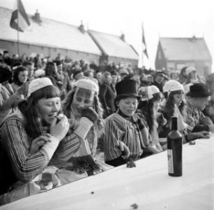 Jonge bruiloftsgasten in klederdracht zitten buiten aan lange tafels Achter hen, Bestanddeelnr 254-1941