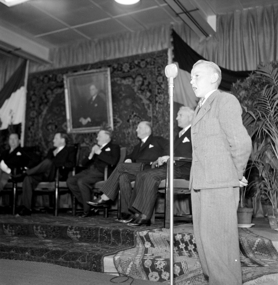 Jongen staand voor een microfoon, op de achtergrond een groep mannen zittend on…, Bestanddeelnr 255-8531