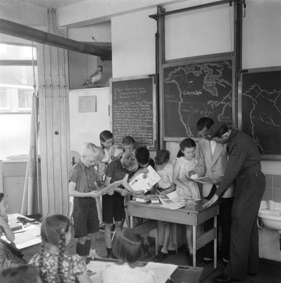 Kinderen verdringen zich rond de tafel van de onderwijzer, naas de onderwijzer , Bestanddeelnr 900-4313