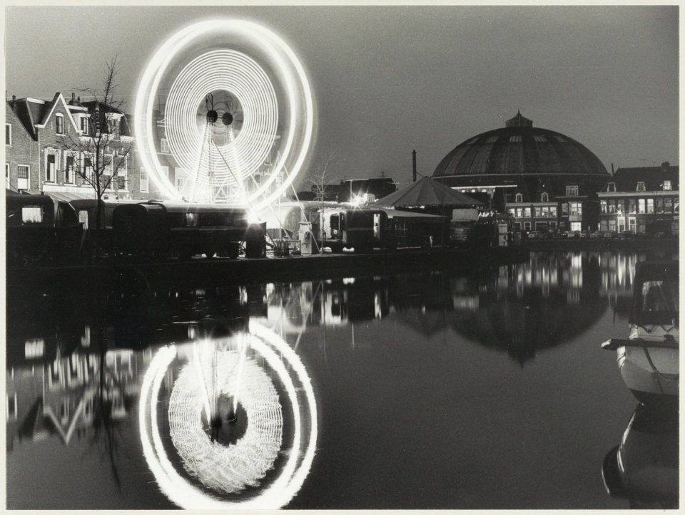 Kermis op het Donkere Spaarne met avondverlichting en rechts de koepelgevangenis. NL-HlmNHA 54011852