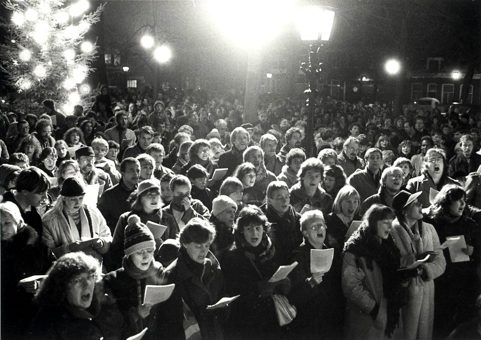 Kerstnacht, samenzang op het Nieuwe Kerksplein, tijdens de kerstnacht. Aangekocht in 1983 van fotograaf C. de Boer. - Negatiefnummer 21457 k 12. - Gepubliceerd in het Haarlems Dagblad van 28, NL-HlmNHA 54013181