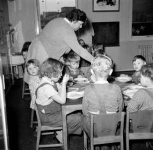 Kleuters zitten aan tafeltjes te eten onder begeleiding van de leidsters, Bestanddeelnr 252-9090
