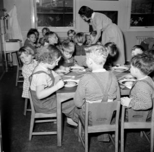 Kleuters zitten aan tafeltjes te eten onder begeleiding van de leidsters, Bestanddeelnr 252-9093