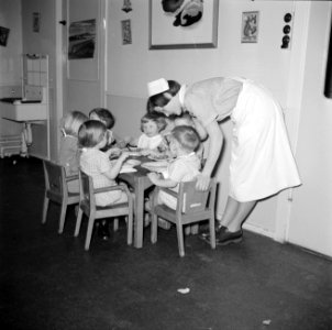 Kleuters zitten aan tafeltjes te eten onder begeleiding van de leidsters, Bestanddeelnr 252-9089