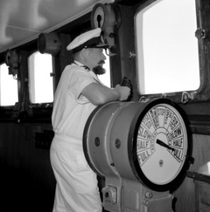 Kapitein in wit uniform met verrekijker in de stuurhut staande bij de scheepstel, Bestanddeelnr 255-4955