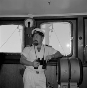 Kapitein in wit uniform met verrekijker in de stuurhut staande bij de scheepstel, Bestanddeelnr 255-4953